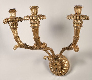 Kinkiet z drewna złoconego, z niewielkim trzonem w kształcie rozety ujętej w trzy liście akantu, z której wyrastają trzy wygięte esowato ramiona świecznika, z okrągłymi profitkami  ze stylizowanych liści i tulejkami na świece utworzonymi z liści ułożonych pionowo.