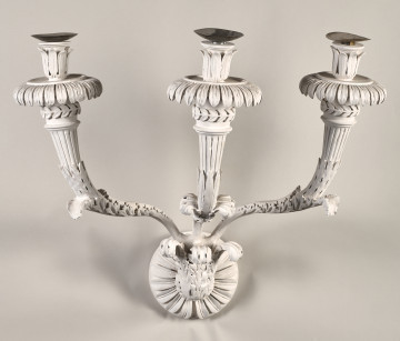 Kinkiet z drewna malowanego na kolor biały, z niewielkim trzonem w kształcie rozety ujętej w trzy liście akantu, z której wyrastają trzy wygięte esowato ramiona świecznika, z okrągłymi profitkami  ze stylizowanych liści i tulejkami na świece utworzonymi z liści ułożonych pionowo.