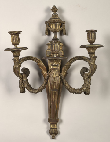 Kinkiet ścienny z wazą z kwiatami w górnej części posiada 3 esowato wygięte ramiona zakończone profitkami i tulejkami na świece, zdobione są stylizowanymi liśćmi akantu i motywem perełek. Przez ramiona przewieszona jest girlanda z liśćmi i dębowymi i żołędziami.