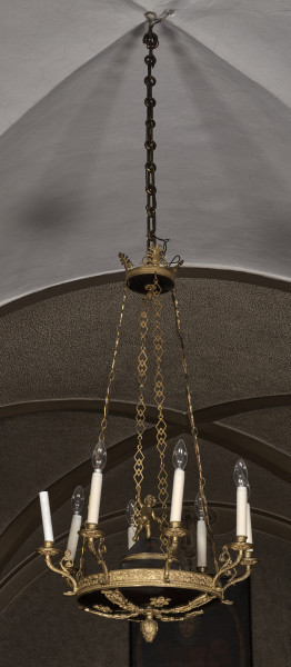 Świecznik zawieszony na czterech łańcuchach, górą schodzących promieniście do zwieńczenia w kształcie obręczy z palmetowymi sterczynami. Korpus świecznika w kształcie płaskiej misy, od góry zamkniętej stożkową pokrywą. Na pokrywie umieszczono postać amorka z podniesioną ręką. Ujęcie na tle białego sklepienia.