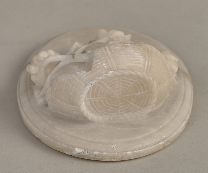 Alabastrowy przycisk do papieru ma kształt owalnego medalionu. Ozdobiony jest półplastyczną kompozycją z kwiatów, gałązek winorośli i owoców umieszczonych w koszu