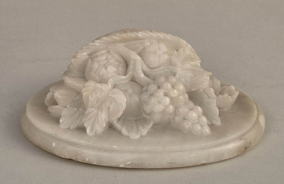 Alabastrowy przycisk do papieru ma kształt owalnego medalionu. Ozdobiony jest półplastyczną kompozycją z kwiatów, gałązek winorośli i owoców umieszczonych w koszu