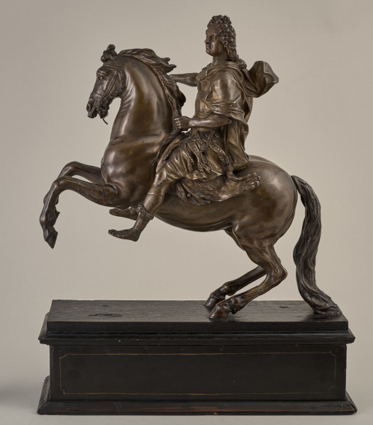 Brązowy posążek na drewnianym postumencie przedstawia Ludwika XIV siedzącego na wspiętym koniu, w stroju i pozie rzymskiego imperatora, w peruce na głowie.