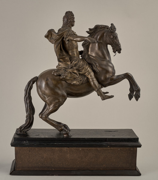 Brązowy posążek na drewnianym postumencie przedstawia Ludwika XIV siedzącego na wspiętym koniu, w stroju i pozie rzymskiego imperatora, w peruce na głowie.