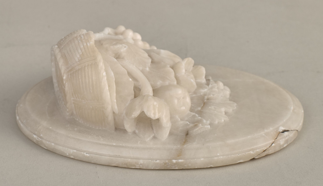 Alabastrowy przycisk do papieru ma kształt owalnego medalionu. Ozdobiony jest półplastyczną kompozycją z kwiatów, gałązek winorośli i owoców umieszczonych w koszu