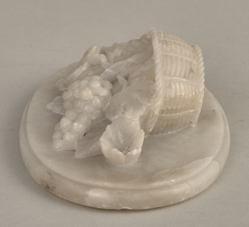 Alabastrowy przycisk do papieru ma kształt owalnego medalionu. Ozdobiony jest półplastyczną kompozycją z kwiatów, gałązek winorośli i owoców umieszczonych w koszu