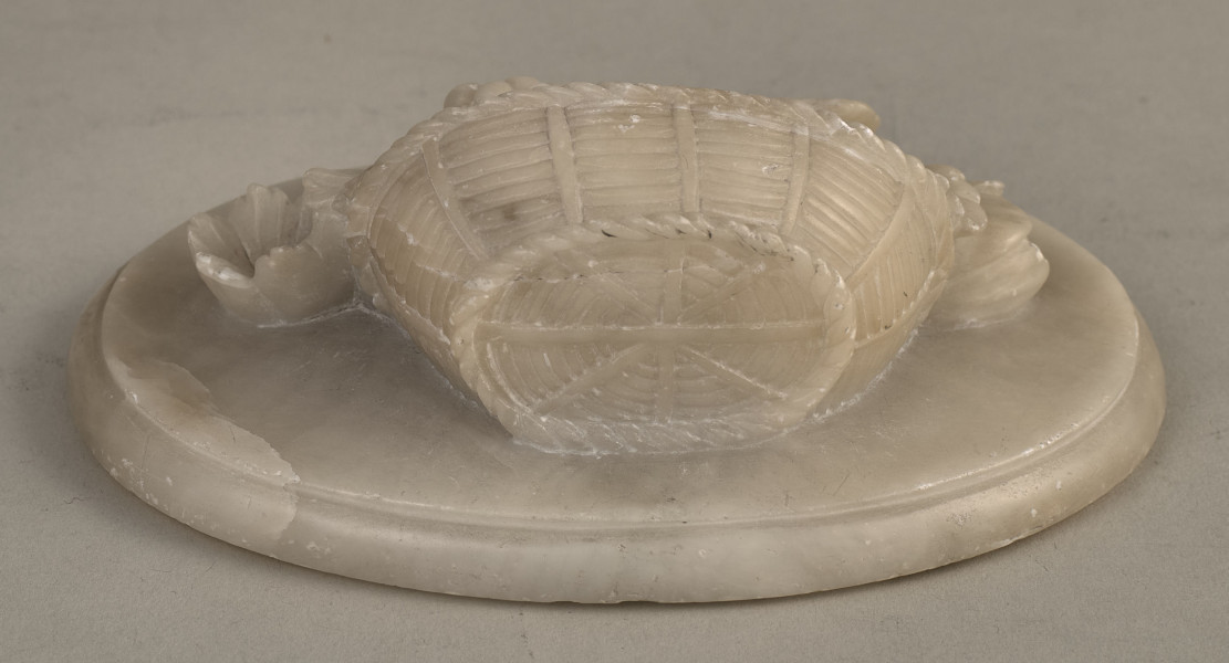 Alabastrowy przycisk do papieru ma kształt owalnego medalionu. Ozdobiony jest półplastyczną kompozycją z kwiatów, gałązek winorośli i owoców umieszczonych w koszu.