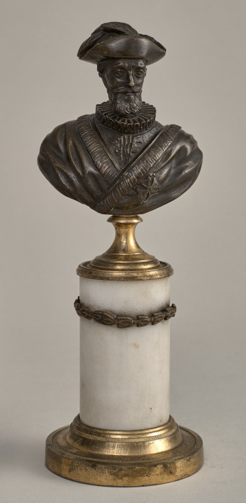 Henryk IV. Brązowe, miniaturowe popiersie przedstawia starszego mężczyznę o szczupłej twarzy, wydatnym nosie, z wąsami i brodą. W kapeluszu z piórami. Ubrany jest w kaftan z wąską krezą i w płaszcz lamowany futrem, na którym widnieje krzyż orderowy. Popiersie ustawione jest na podstawie złożonej z mosiężnego cokoliku, marmurowego trzonu ujętego metalowym łańcuszkiem i mosiężnej bazy. Przód