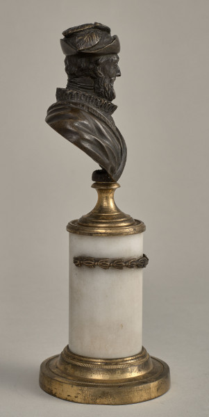 Henryk IV. Brązowe, miniaturowe popiersie przedstawia starszego mężczyznę o szczupłej twarzy, wydatnym nosie, z wąsami i brodą. W kapeluszu z piórami. Ubrany jest w kaftan z wąską krezą i w płaszcz lamowany futrem, na którym widnieje krzyż orderowy. Popiersie ustawione jest na podstawie złożonej z mosiężnego cokoliku, marmurowego trzonu ujętego metalowym łańcuszkiem i mosiężnej bazy. Bok prawy