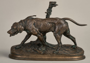 Ogar. Posążek z brązu. Ogar ukazany został w wykroku do przodu przednią prawą łapą, ze spuszczonym łbem i wysuniętym językiem; w obroży ze smyczą przywiązaną do pnia drzewa. Podstawa imituje naturalne podłoże ziemi. Bok lewy