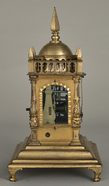 Zegar wieżyczkowy. Zegar złocony w kształcie małej wieży zegarowej zwieńczony galeryjką arkadową z czterema sterczynkami na narożach oraz kopulastym bębnem ze sterczyną. Pod arkadką duże okno ukazujące wewnętrzny mechanizm.