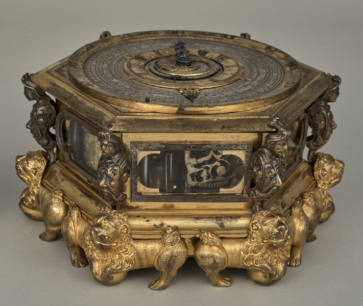 Zegar kaflak z tarczą poziomą. Oprawę zegara stanowi sześcioboczna puszka, której narożniki dekorowane są wąsatymi rycerzami, natomiast podobne są maszkarony zdobiące piersi tych herm i nóżki w kształcie figurek toczących kule lwów o dekoracyjnych ogonach i rozdwajających się tułowiach. Oszklone okienka są obramione na przemian grawerowaną opaską prostokątną oraz połączeniem prostokąta i półkola. Całość zegara została wykonana z brązu złoconego