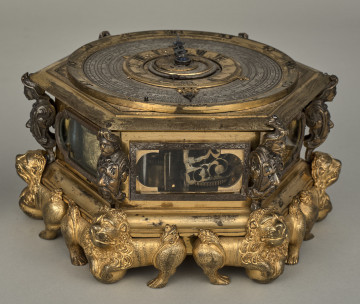 Zegar kaflak z tarczą poziomą. Oprawę zegara stanowi sześcioboczna puszka, której narożniki dekorowane są wąsatymi rycerzami, natomiast podobne są maszkarony zdobiące piersi tych herm i nóżki w kształcie figurek toczących kule lwów o dekoracyjnych ogonach i rozdwajających się tułowiach. Oszklone okienka są obramione na przemian grawerowaną opaską prostokątną oraz połączeniem prostokąta i półkola. Całość zegara została wykonana z brązu złoconego