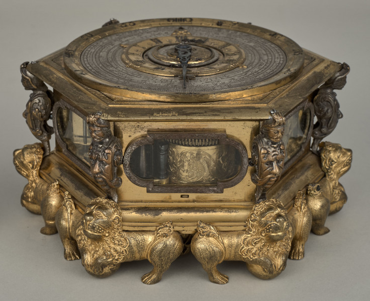 Zegar kaflak z tarczą poziomą. Oprawę zegara stanowi sześcioboczna puszka, której narożniki dekorowane są wąsatymi rycerzami, natomiast podobne są maszkarony zdobiące piersi tych herm i nóżki w kształcie figurek toczących kule lwów o dekoracyjnych ogonach i rozdwajających się tułowiach. Oszklone okienka są obramione na przemian grawerowaną opaską prostokątną oraz połączeniem prostokąta i półkola. Całość zegara została wykonana z brązu złoconego