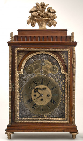 Zegar kominkowy w drewnianej obudowie ze złoceniami. Przednie drzwiczki szklone, ukazujące mosiężną tarczę zegarową z rzymskimi cyframi. U góry dwie dodatkowe tarcze. Wnętrze dekorowane srebrną blachą z repusowanymi wzorami roślinnymi. Na szczycie skrzynki złoty wazon z kwiatami.