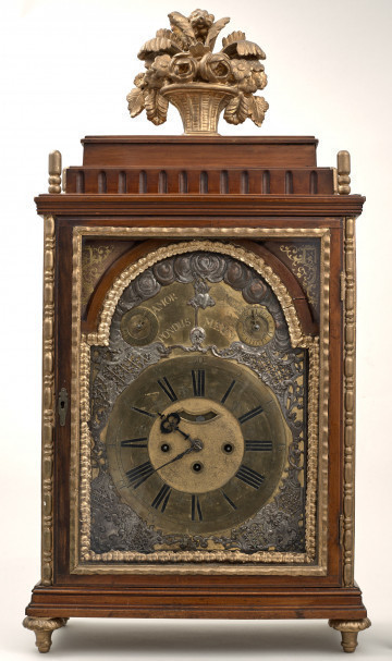 Zegar kominkowy w drewnianej obudowie ze złoceniami. Przednie drzwiczki szklone, ukazujące mosiężną tarczę zegarową z rzymskimi cyframi. U góry dwie dodatkowe tarcze. Wnętrze dekorowane srebrną blachą z repusowanymi wzorami roślinnymi. Na szczycie skrzynki złoty wazon z kwiatami.