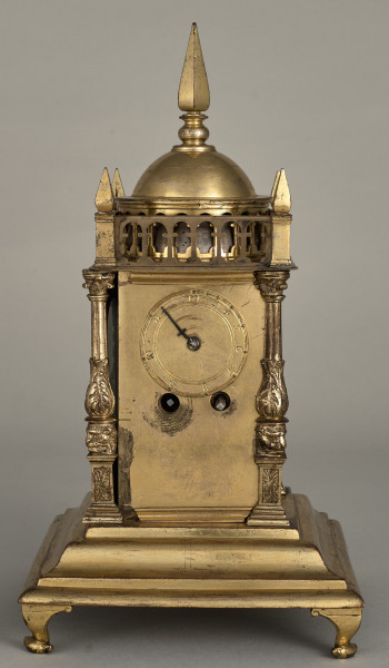 Zegar wieżyczkowy. Zegar złocony w kształcie małej wieży zegarowej zwieńczony galeryjką arkadową z czterema sterczynkami na narożach oraz kopulastym bębnem ze sterczyną. Pod arkadką mniejsza tarcza z jedną wskazówką i otwory do nakręcania zegara.