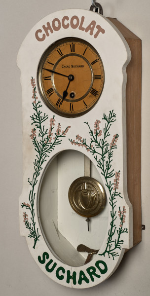 Zegar wiszący. ﻿Bardzo oryginalny zegar reklamowy firmy Suchard. Ścianka frontowa skrzynki zegarowej zdobiona jest motywem lawendy i napisem CHOCOLAT SUCHARD. W ściance znajdują się dwa otwory, z których górny pozwala obserwować białą, emaliowaną tarczę zegara z wykonanymi w kolorze czarnym cyframi rzymskimi. Poniżej znajduje się owalny otwór, przez który widoczna jest praca wahadła. Przód 3/4
