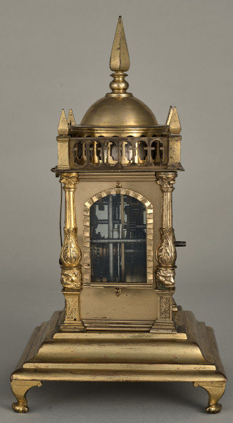 Zegar wieżyczkowy. Zegar złocony w kształcie małej wieży zegarowej zwieńczony galeryjką arkadową z czterema sterczynkami na narożach oraz kopulastym bębnem ze sterczyną. Pod arkadką duże okno ukazujące wewnętrzny mechanizm.