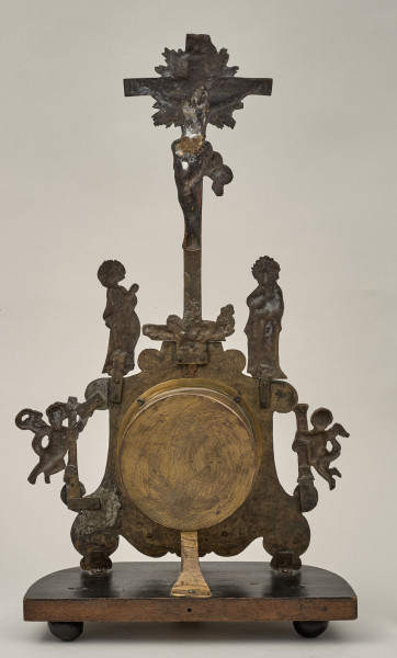 Zegar pasyjka. Mosiężna obudowa mechanizmu na płaskim kartuszu. Na szczycie scena Ukrzyżowania Chrystusa z postaciami towarzyszącymi - Marią i świętym Janem Apostołem. Po bokach tarczy aniołki. Zegar ukazany od tyłu. Postacie i dekoracje widoczne w negatywie.