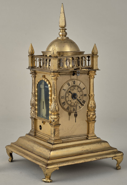 Zegar wieżyczkowy. Zegar złocony w kształcie małej wieży zegarowej zwieńczony galeryjką arkadową z czterema sterczynkami na narożach oraz kopulastym bębnem ze sterczyną. Pod arkadką duża tarcza zegarowa. Ujęcie  3/4 z widoczną tarczą oraz oknem.