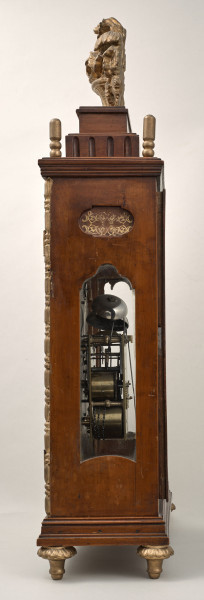 Zegar kominkowy w drewnianej obudowie ze złoceniami. Boczna ścianka z otworem szklonym ukazującym mechanizm.