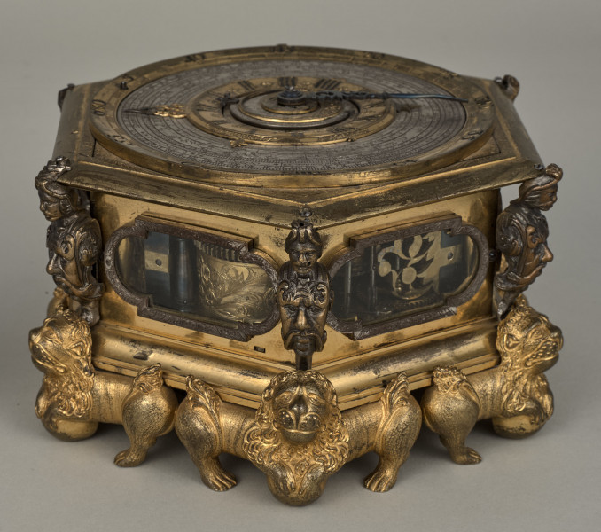 Zegar kaflak z tarczą poziomą. Oprawę zegara stanowi sześcioboczna puszka, której narożniki dekorowane są wąsatymi rycerzami, natomiast podobne są maszkarony zdobiące piersi tych herm i nóżki w kształcie figurek toczących kule lwów o dekoracyjnych ogonach i rozdwajających się tułowiach. Oszklone okienka są obramione na przemian grawerowaną opaską prostokątną oraz połączeniem prostokąta i półkola. Całość zegara została wykonana z brązu złoconego