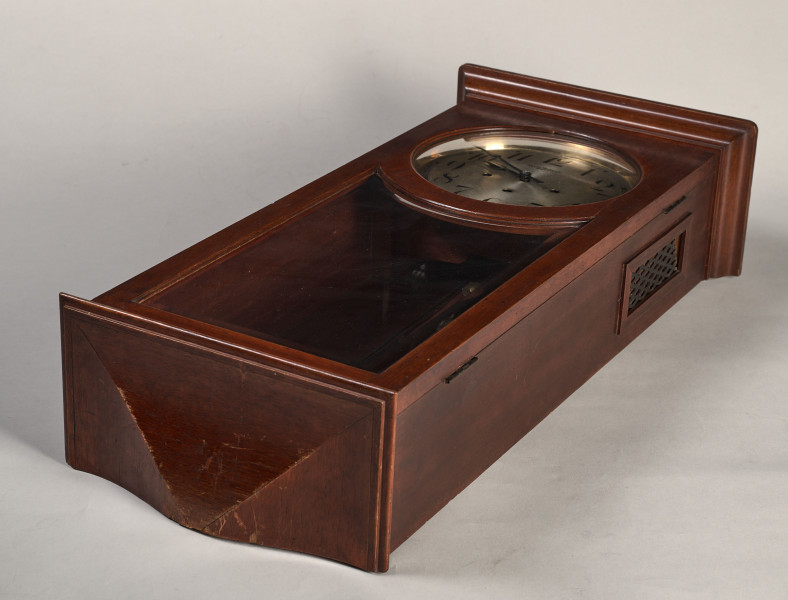 Zegar szafkowy, wiszący ma kształt prostopadłościanu z gzymsem i profilowanym cokołem. W dolnej części jest przeszklony. Widok leżącego na tylnej ściance zegara.