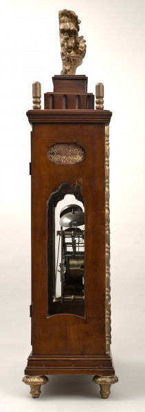 Zegar kominkowy w drewnianej obudowie ze złoceniami. Boczna ścianka z otworem szklonym ukazującym mechanizm.