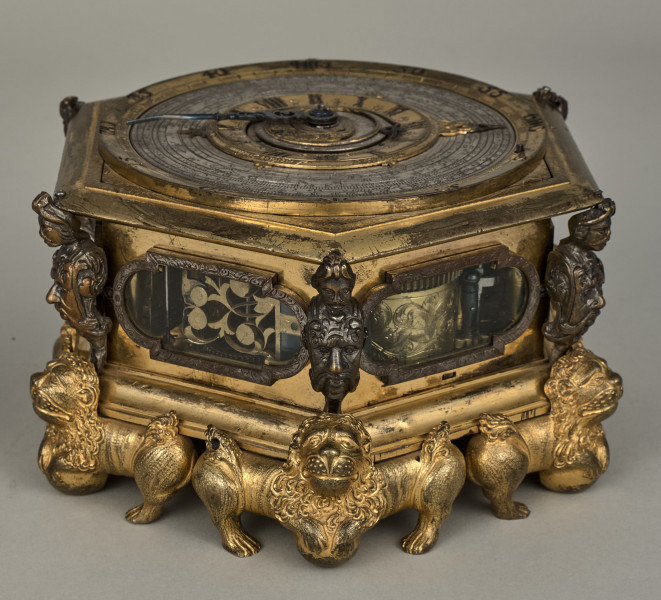 Zegar kaflak z tarczą poziomą. Oprawę zegara stanowi sześcioboczna puszka, której narożniki dekorowane są wąsatymi rycerzami, natomiast podobne są maszkarony zdobiące piersi tych herm i nóżki w kształcie figurek toczących kule lwów o dekoracyjnych ogonach i rozdwajających się tułowiach. Oszklone okienka są obramione na przemian grawerowaną opaską prostokątną oraz połączeniem prostokąta i półkola. Całość zegara została wykonana z brązu złoconego