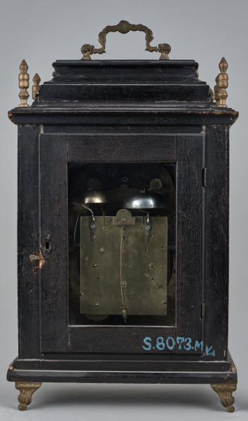 Zegar szafkowy z uchwytem górnym do przenoszenia. Prostopadłościenna obudowa drewniana, czerniona, złocona, posiada aplikacje mosiężne. Ścianka tylna z otworem, przez który widać mechanizm.