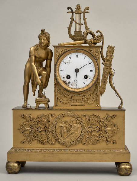 Przód złoconego zegara kominkowego. Podstawa na kulistych nóżkach, zdobiona ornamentem roślinnym z centralnie umieszczonym medalionem z postaciami ludzkimi. Na postumencie postać ludzka, pełnoplastyczna obok tarcza zegarowa w złotej obudowie. Po prawej stronie łuk z kołczanem. Na obudowie stoi lira.