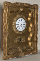 Zegar wiszący, złocony w formie przeszklonej otwieranej szafki w ramie ozdobnej. Biała tarcza zegarowa ozdobiona wieńcem rzeźbionym. Poniżej tarczy otwór. Ujęcie 3/4