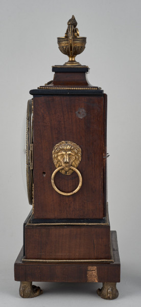 Bok zegara szafkowego. Obudowa drewniana, ciemna na zoomorficznych nóżkach. W środkowej części uchwyt z formie lwiej głowy. U samej góry waza.