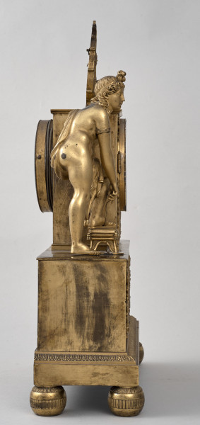 Boczna ścianka zegara kominkowego, złoconego. Podstawa na kulistych nóżkach. Na podstawie postać pełnoplastyczna, a nad nią widoczna lira