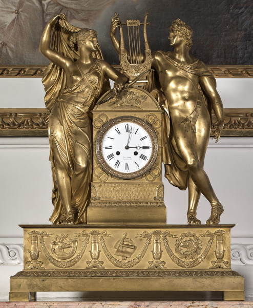 Zegar kominkowy wykonany z brązu złoconego, którego dekoracyjna obudowa przedstawia postaci mitologiczne Dianę trzymającą łuk i Apollina z lirą opartych na prostopadłościennej obudowie werku, która została umieszczona na postumencie w typie sarkofagu, wspartym na czterech prostopadłościennych nóżkach. Tarcza biała, emaliowana z cyframi rzymskimi dla godzin i arabskimi dla minut została wkomponowana w mosiężną płytkę.