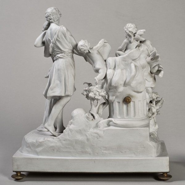 ﻿Prezentowany zegar został wykonany z biskwitowej porcelany twardej, w pełnoplastycznej oprawie rzeźby figuralnej. Tematem jest para młodych pasterzy zapatrzonych w siebie ujęta w ruchu tanecznym. Kierują się w stronę ściętej kolumny, a na jej szczycie siedzą trzy uskrzydlone amorki, z których jeden trzyma w dłoniach róg obfitości. Ponadto u stóp pasterzy znajduje się, laska, kapelusz, dzban i koszyk. Dopełnieniem sceny są dwa gołąbki i konary krzewów. Stroje postaci nawiązują do antyku. Tył