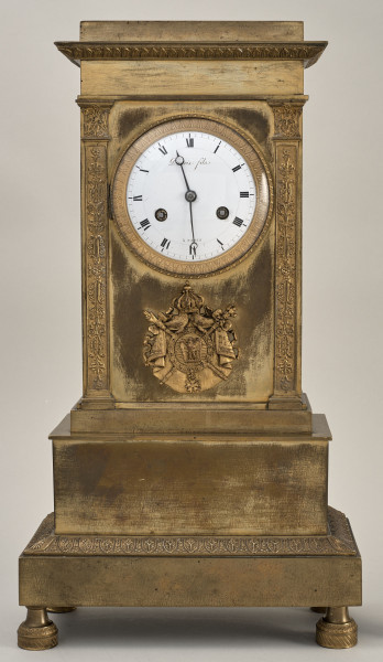 Przód zegara kominkowego w formie architektonicznej w kolorze złotym z białą tarczą zegarową i przedstawieniem herbowym pod nią