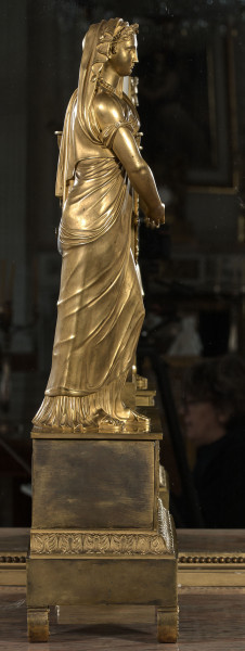 Bok zegara kominkowego, złoconego, o dwóch poziomach kompozycji. Podstawa na nóżkach, zdobiona pasem kimationu. Powyżej pełnoplastyczna postać kobieca.