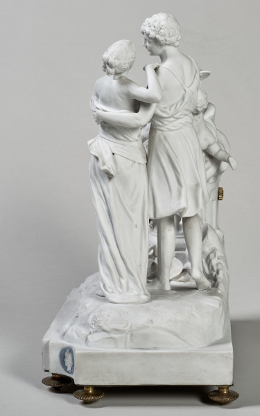 ﻿Prezentowany zegar został wykonany z biskwitowej porcelany twardej, w pełnoplastycznej oprawie rzeźby figuralnej. Tematem jest para młodych pasterzy zapatrzonych w siebie ujęta w ruchu tanecznym. Kierują się w stronę ściętej kolumny, a na jej szczycie siedzą trzy uskrzydlone amorki, z których jeden trzyma w dłoniach róg obfitości. Ponadto u stóp pasterzy znajduje się, laska, kapelusz, dzban i koszyk. Dopełnieniem sceny są dwa gołąbki i konary krzewów. Stroje postaci nawiązują do antyku. Bok lewy