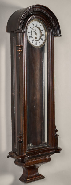 Zegar szafkowy, wiszący w wąskiej, ciemnej, drewnianej obudowie u góry zakończonej półokrągłym daszkiem. Tarcza zegarowa biała znajduje się u samej góry. Ujęcie 3/4