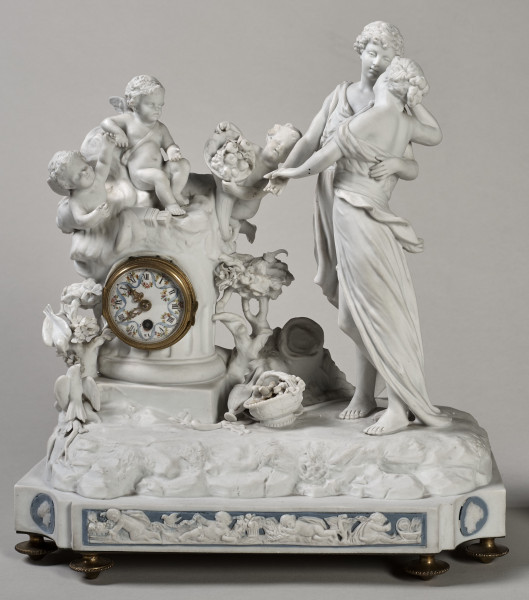 ﻿Prezentowany zegar został wykonany z biskwitowej porcelany twardej, w pełnoplastycznej oprawie rzeźby figuralnej. Tematem jest para młodych pasterzy zapatrzonych w siebie ujęta w ruchu tanecznym. Kierują się w stronę ściętej kolumny, a na jej szczycie siedzą trzy uskrzydlone amorki, z których jeden trzyma w dłoniach róg obfitości. Ponadto u stóp pasterzy znajduje się, laska, kapelusz, dzban i koszyk. Dopełnieniem sceny są dwa gołąbki i konary krzewów. Stroje postaci nawiązują do antyku. Mechanizm zegara został wmontowany w kolumnę. Na białej tarczy cyfry rzymskie wskazujące godziny. Podłoże nawiazuje do ziemi pokrytej roślinnością. Ujęcie frontalne