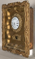 Zegar wiszący, złocony w formie przeszklonej otwieranej szafki w ramie ozdobnej. Biała tarcza zegarowa ozdobiona wieńcem rzeźbionym. Poniżej tarczy otwór. Ujęcie 3/4