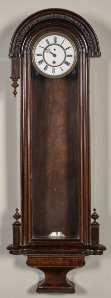 Zegar szafkowy, wiszący w wąskiej, ciemnej, drewnianej obudowie u góry zakończonej półokrągłym daszkiem. Tarcza zegarowa biała znajduje się u samej góry. Ujęcie frontalne