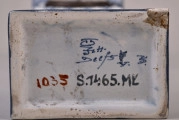 Fragment ceramicznej podstawy zegara z widocznymi znakami producenta oraz numerem inwentarzowym muzeum.