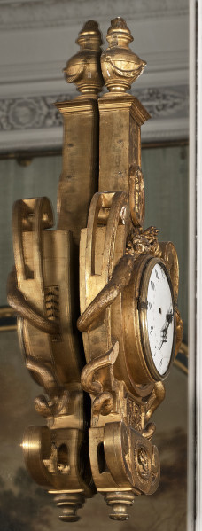 Zegar wiszący na lustrze, drewniany, złocony w kształcie liry z wijącymi wężami wokół ramion, zwieńczony wazonem. Tarcza okrągła z ażurowymi wskazówkami, emaliowana na biało, na niej czarne cyfry arabskie. Bok lewy