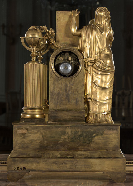 Tył zegara kominkowego, złoconego, o dwóch poziomach kompozycji. Podstawa na nóżkach z pasem kimationu. Powyżej obudowa tarczy zegara flankowana po prawej stronie pełnoplastyczną postacią kobiecą, po lewej kolumną z globusem i ulistnioną gałęzią.