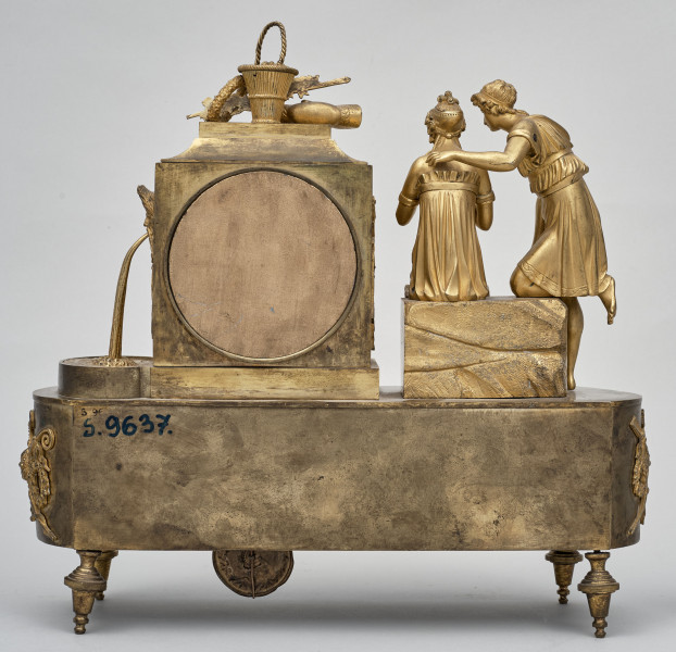 Tył zegara kominkowego. Podstawa na nóżkach. Na podstawie po prawej stronie dwie postacie pełnoplastyczne stojące tyłem. Po lewej obudowa tarczy zegarowej.