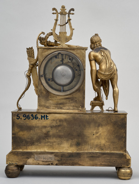 Tył złoconego zegara kominkowego. Podstawa na kulistych nóżkach. Na postumencie postać ludzka, pełnoplastyczna obok obudowa tarczy z otworem na mechanizm. Po lewej stronie łuk z kołczanem. Na obudowie stoi lira.