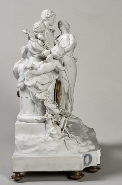 ﻿Prezentowany zegar został wykonany z biskwitowej porcelany twardej, w pełnoplastycznej oprawie rzeźby figuralnej. Tematem jest para młodych pasterzy zapatrzonych w siebie ujęta w ruchu tanecznym. Kierują się w stronę ściętej kolumny, a na jej szczycie siedzą trzy uskrzydlone amorki, z których jeden trzyma w dłoniach róg obfitości. Ponadto u stóp pasterzy znajduje się, laska, kapelusz, dzban i koszyk. Dopełnieniem sceny są dwa gołąbki i konary krzewów. Stroje postaci nawiązują do antyku. Bok prawy